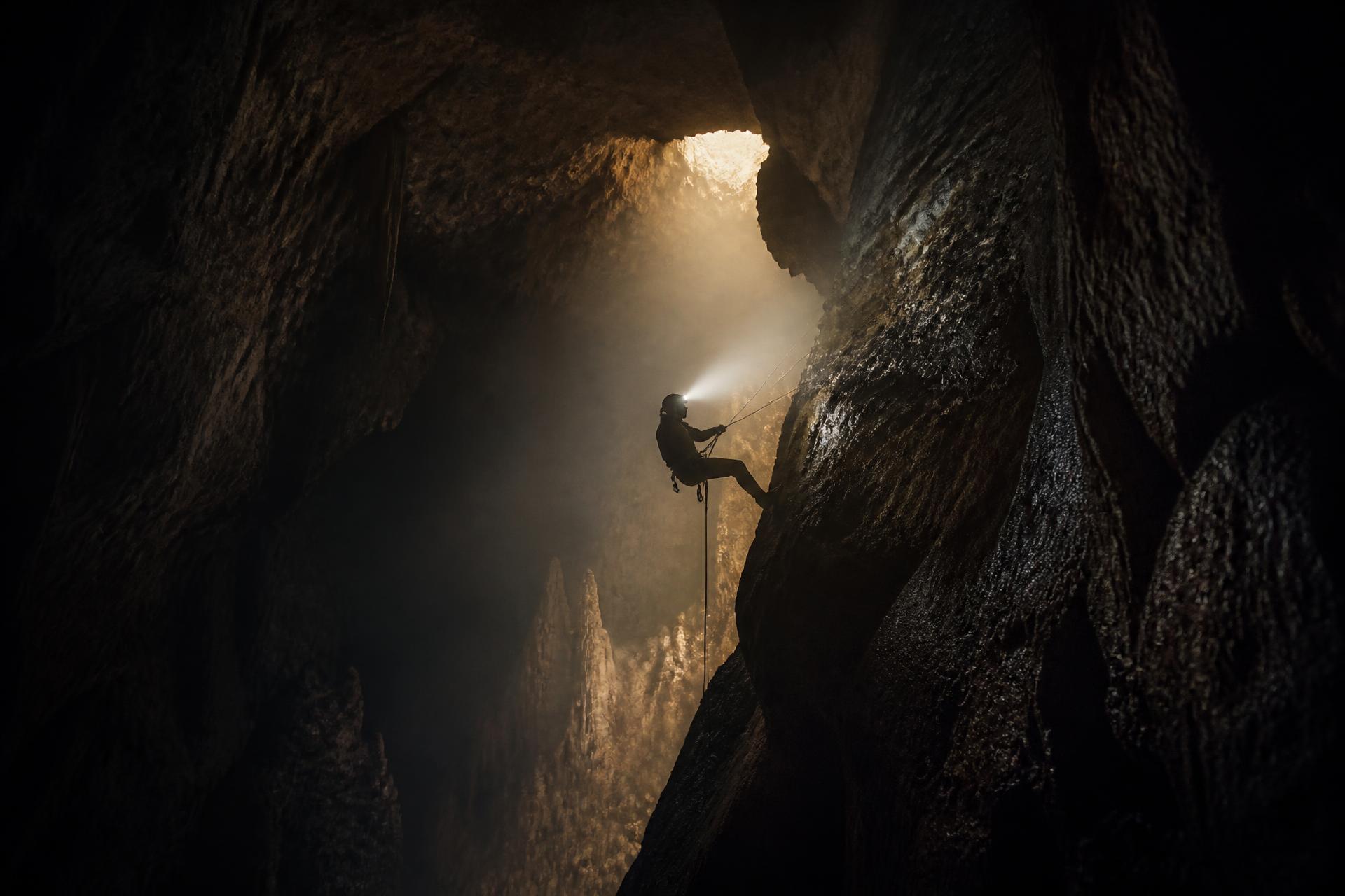 Caver abseiling through a shaft of light inside a limestone cavern